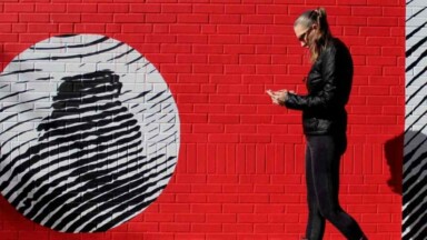 Frau läuft mit Handy in der Hand vor einer roten Wand entlang. Überdimensionaler Fingerabdruck für Digitale Identitäten