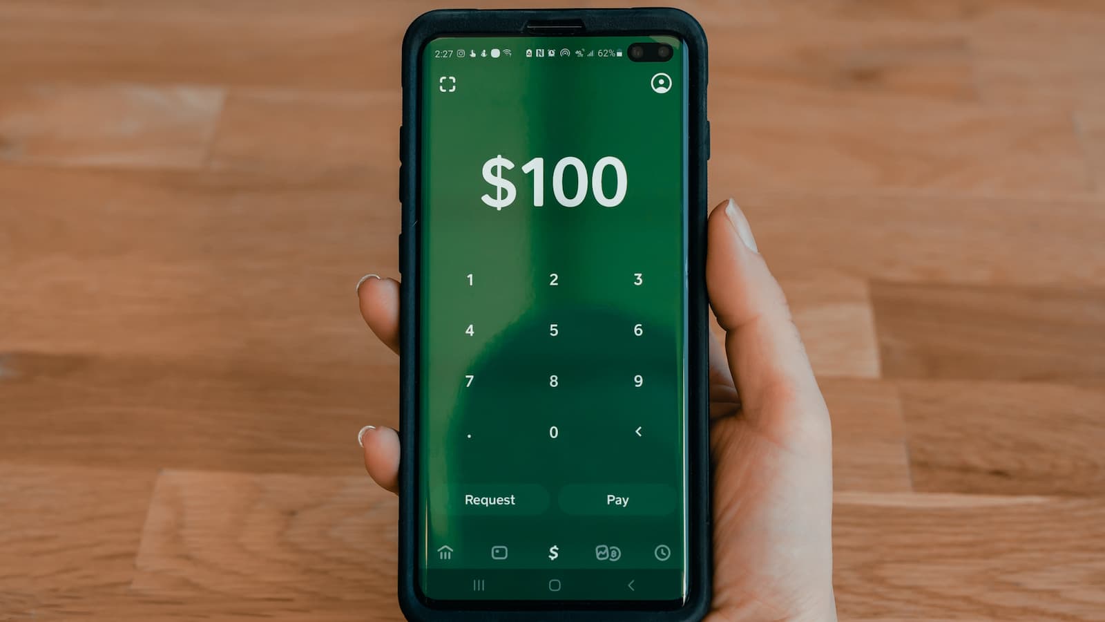 Hand hält Smartphone mit grünem Cash App Hintergrund und 100$ Anzeige, Parkett im Hintergrund