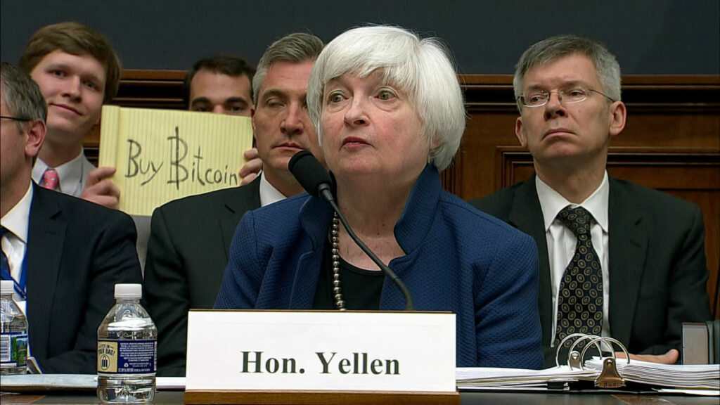 Yellen vor dem US-Kongress. Im Hintergrund hält ein Besucher ein Schild mit dem Spruch „kauft Bitcoin