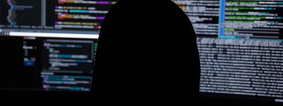 symboldbild: hacker sitzt in dunkler kapuze vor bildschirmen mit programmcode