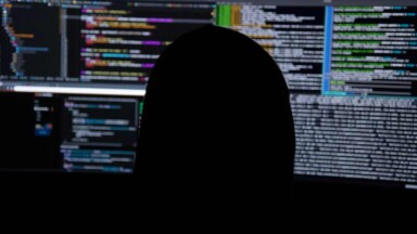 symboldbild: hacker sitzt in dunkler kapuze vor bildschirmen mit programmcode